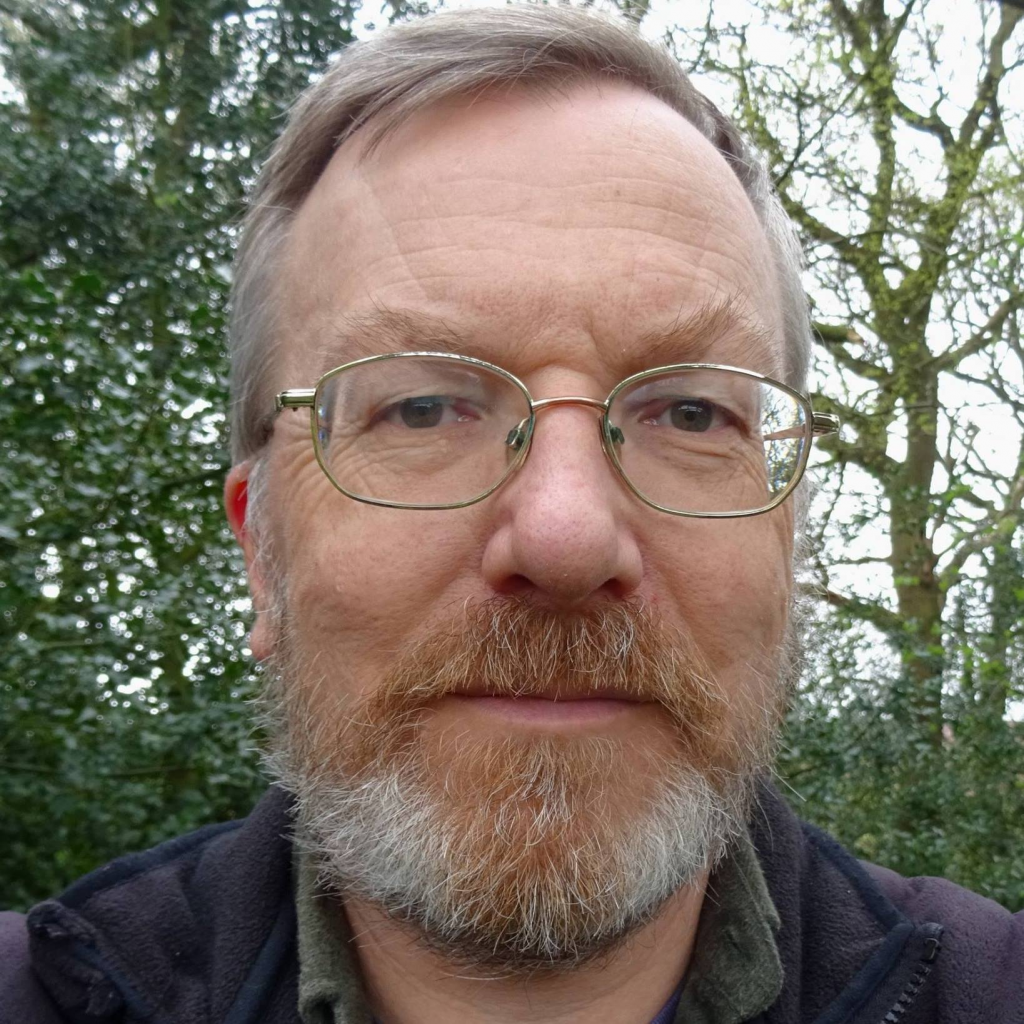 A photograph of Peter Hughes. A white man with a beard and glasses with fair hair standing in front of trees. Peter has a warm expression.