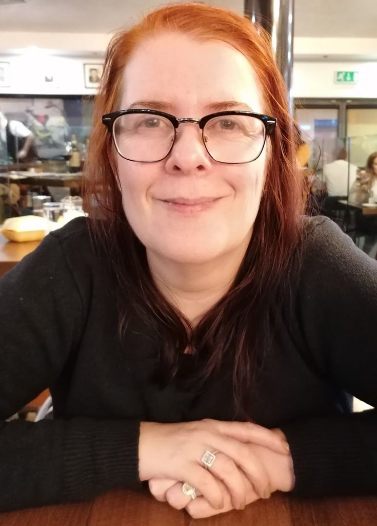 A photograph of Prof. Sam Broadhead. She is white, has red hair, is wearing glasses and is smiling. 